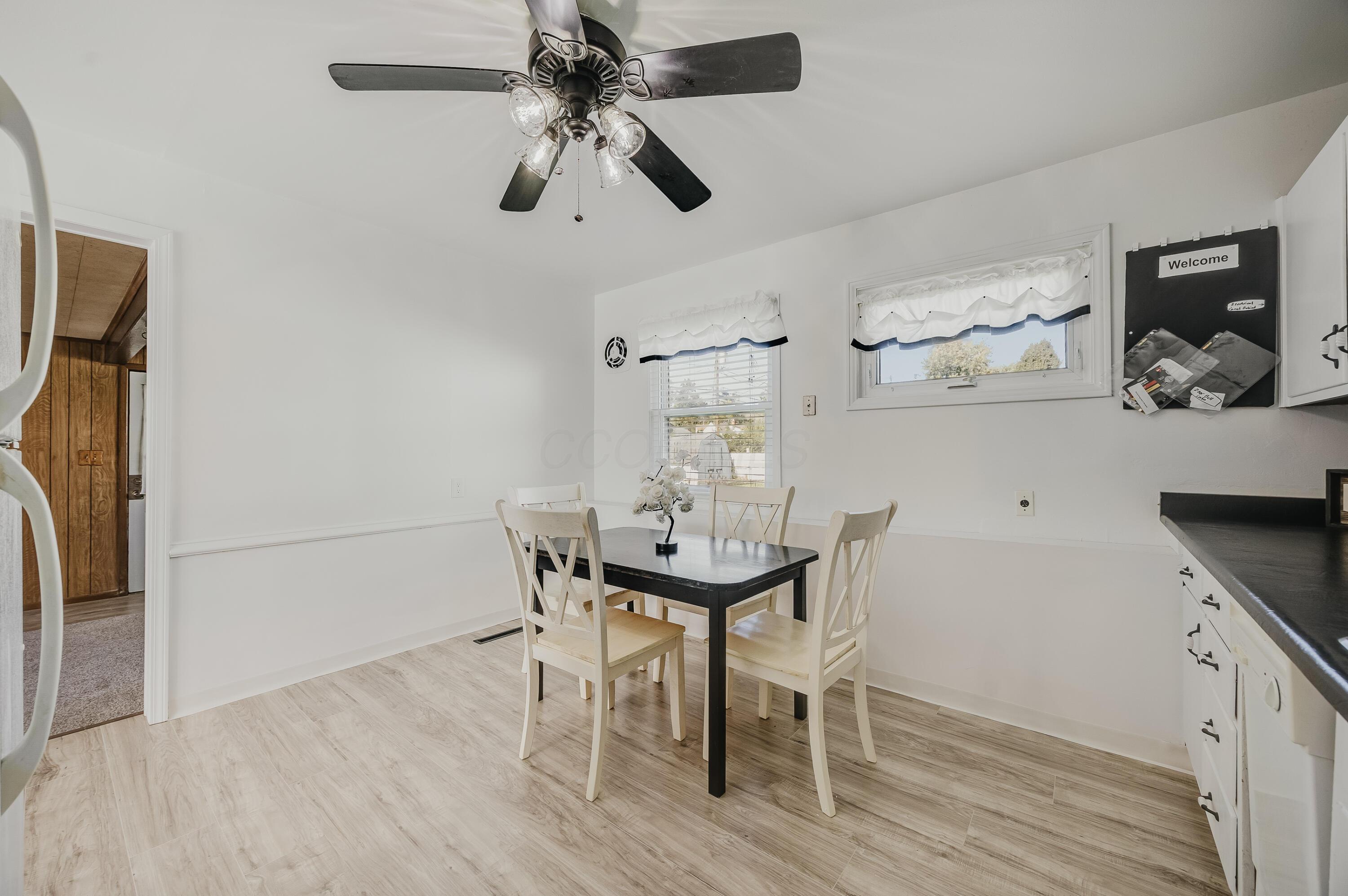 Dining area with a black table, light wood chairs, two curtained windows, and a ceiling fan.