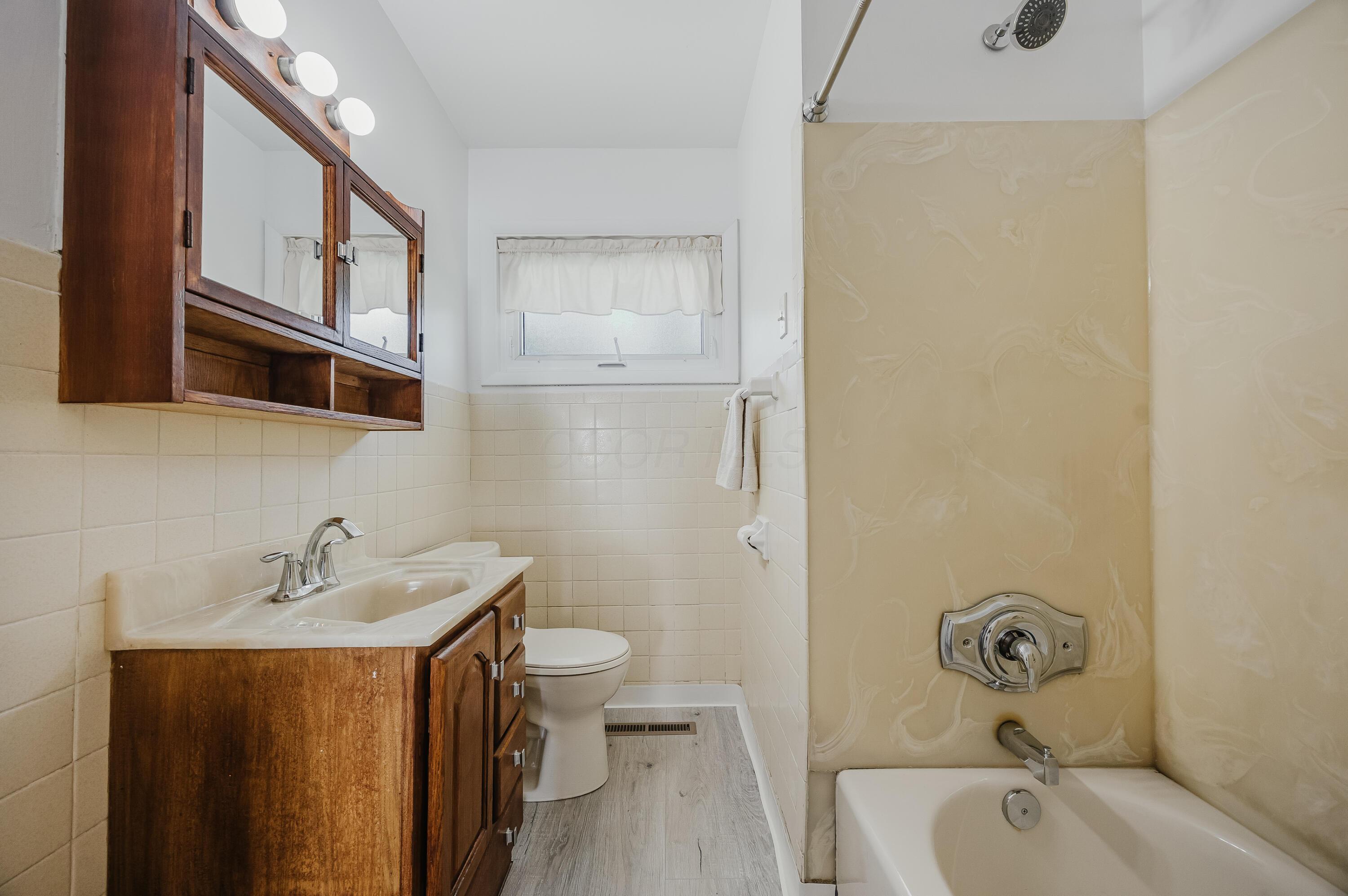Bathroom with a wooden vanity, toilet beneath a window, and bathtub with beige patterned surround.