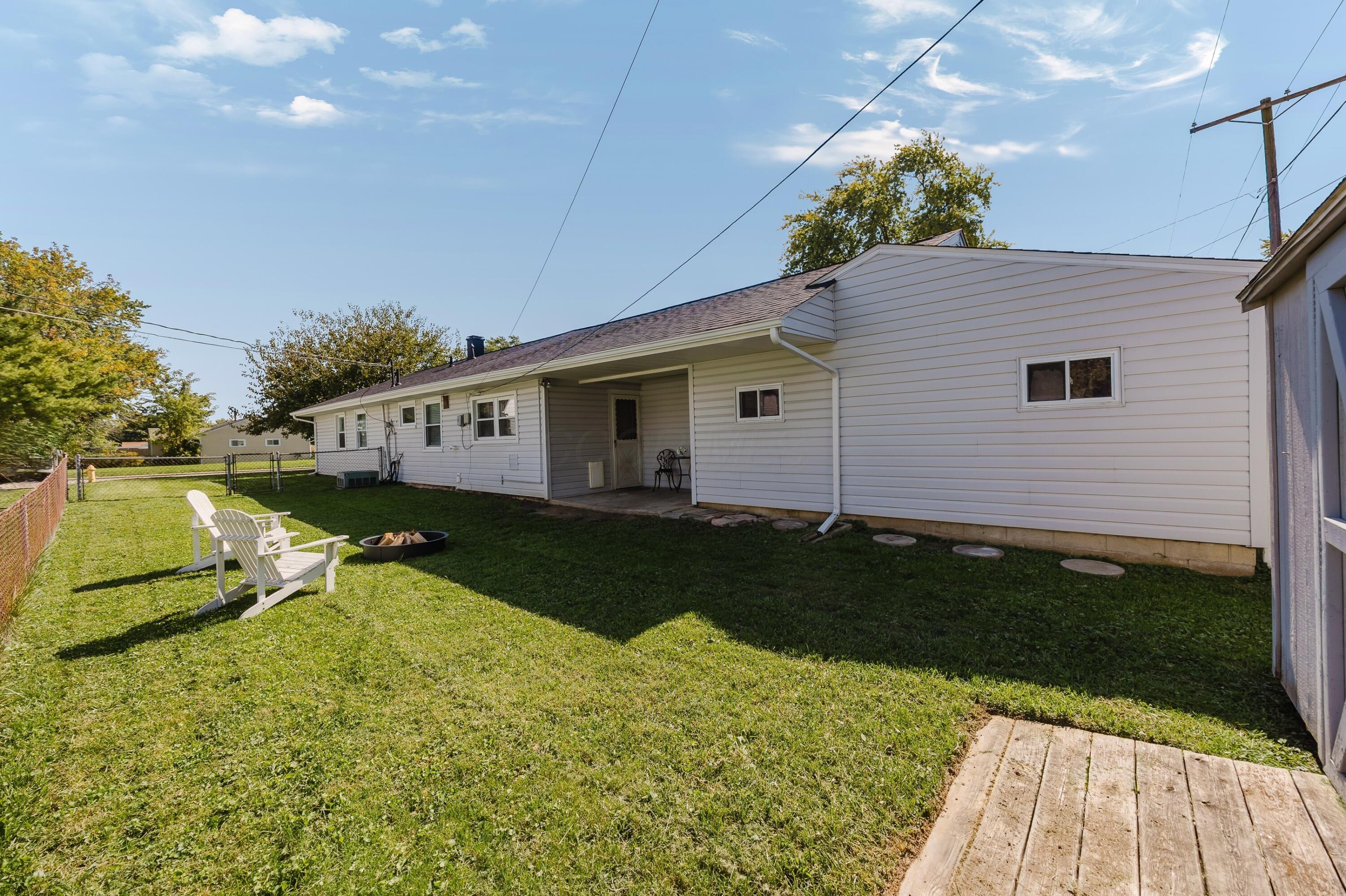Rear of a white-sided home with a covered patio, grassy yard, white chairs, and a fire pit.