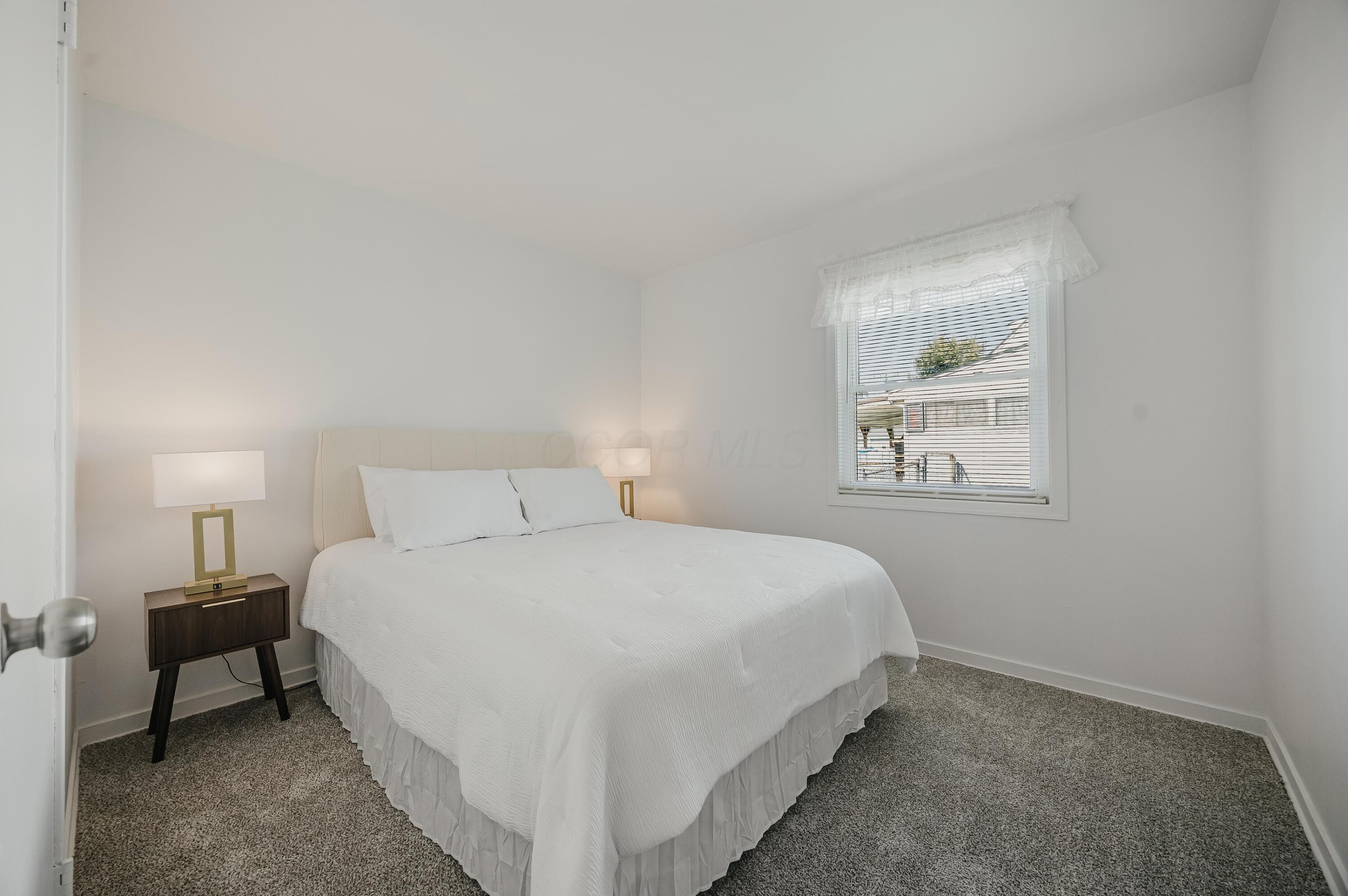 Staged bedroom with a white bed, dark nightstand, gray carpet, and window with blinds.