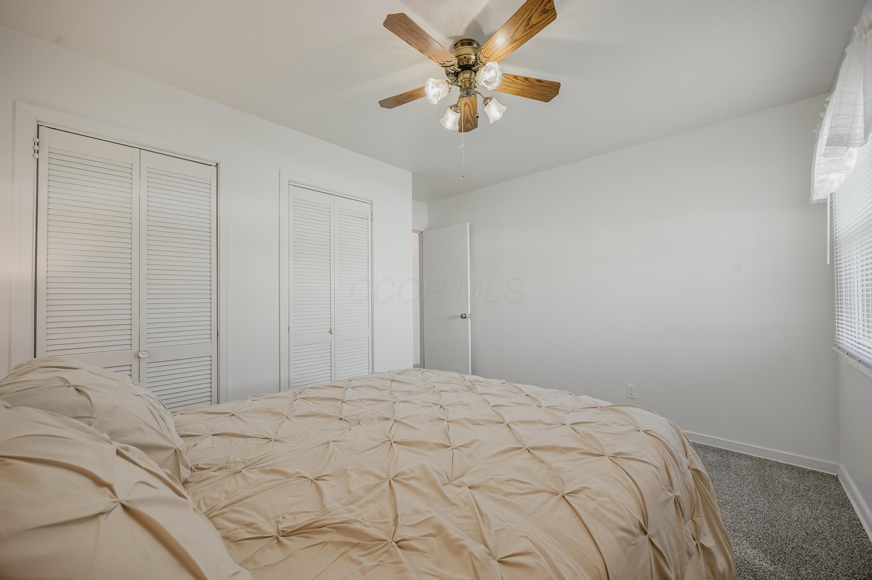 Bedroom with cream bedding, louvered closet doors, and a wood-blade ceiling fan.