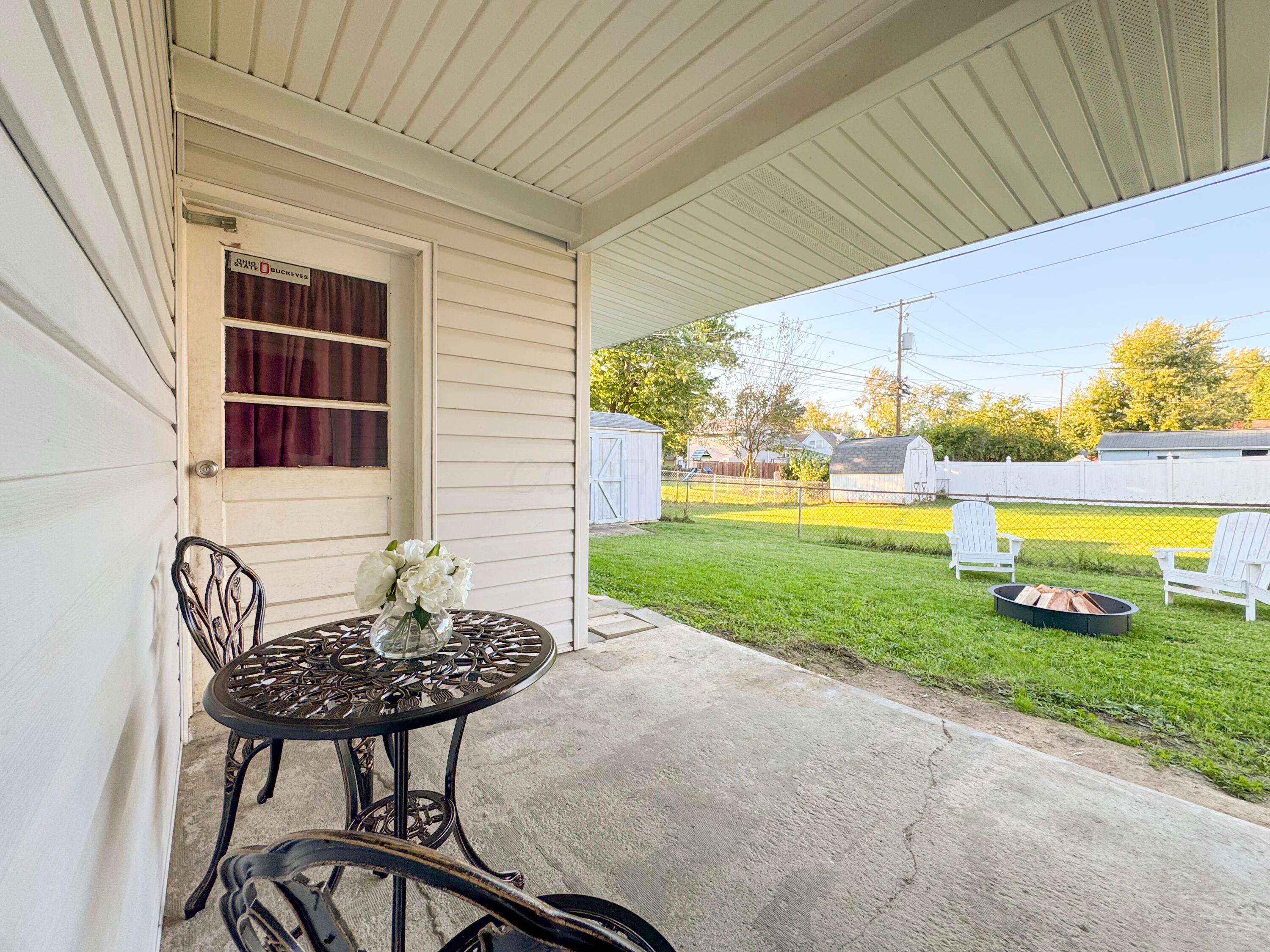 Covered patio with black metal furniture overlooking a fenced backyard with Adirondack chairs and a fire pit.