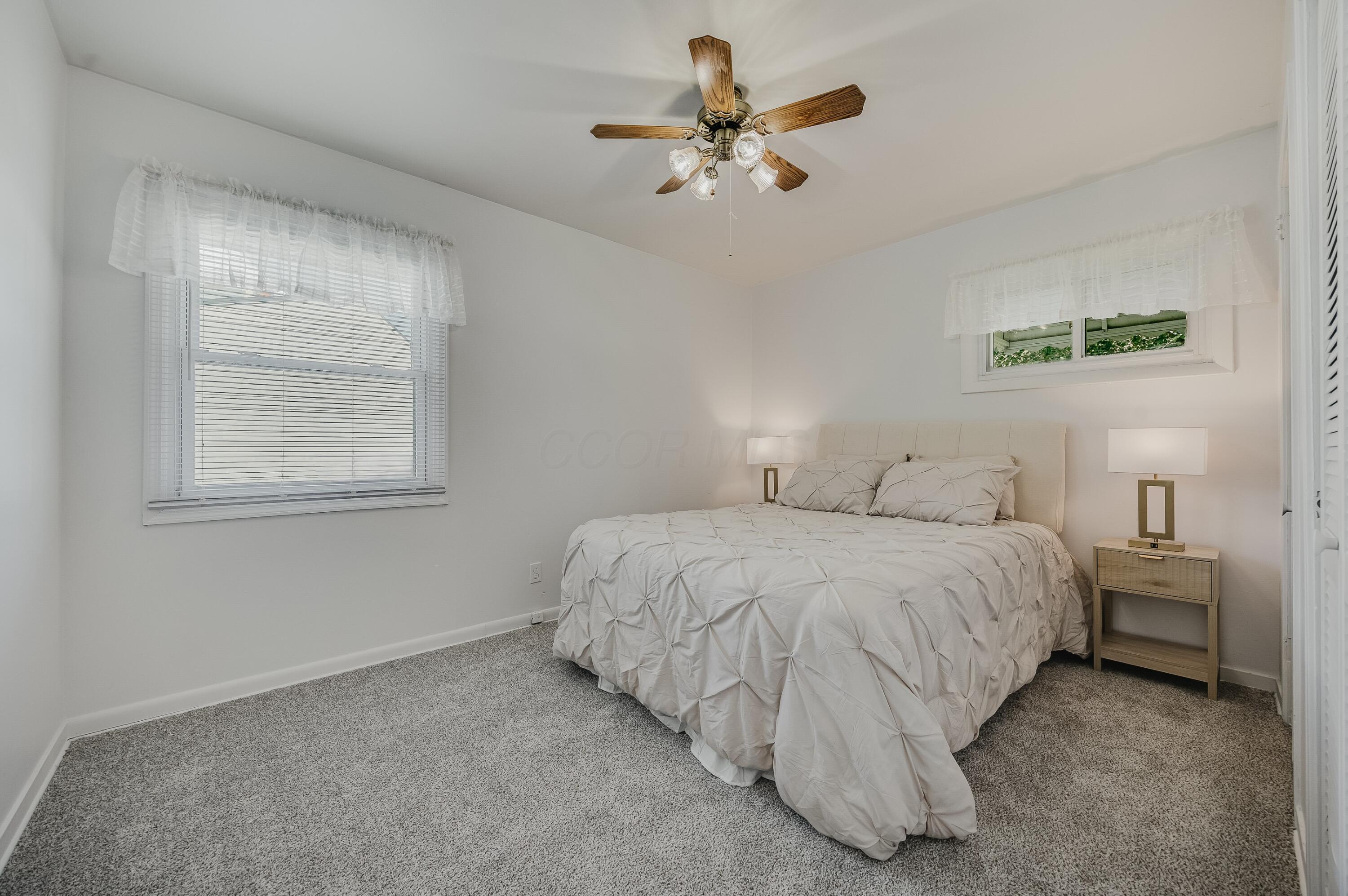 Furnished bedroom with a large bed, gray carpet, two windows, and a wooden ceiling fan.