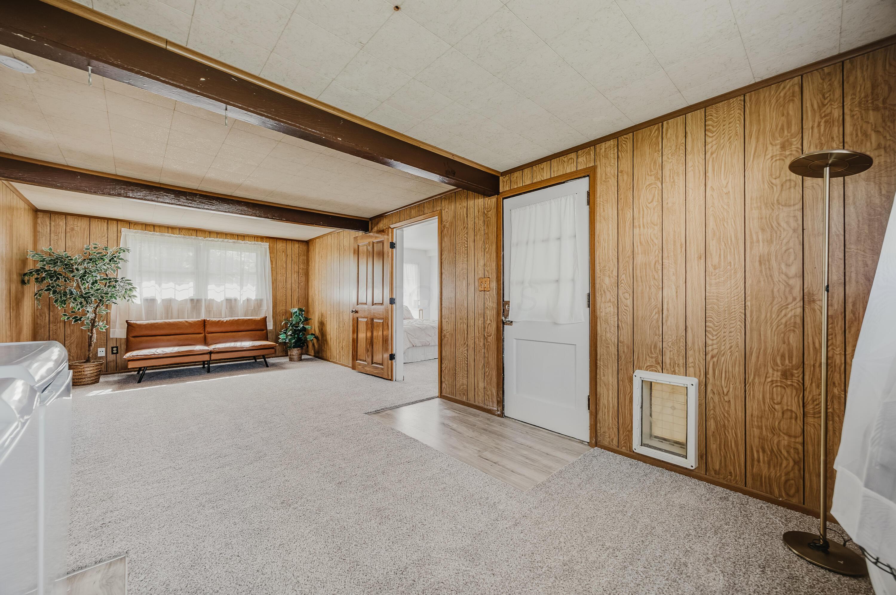 Living room with wood-paneled walls, exposed ceiling beams, and a brown sofa beneath a curtained window.