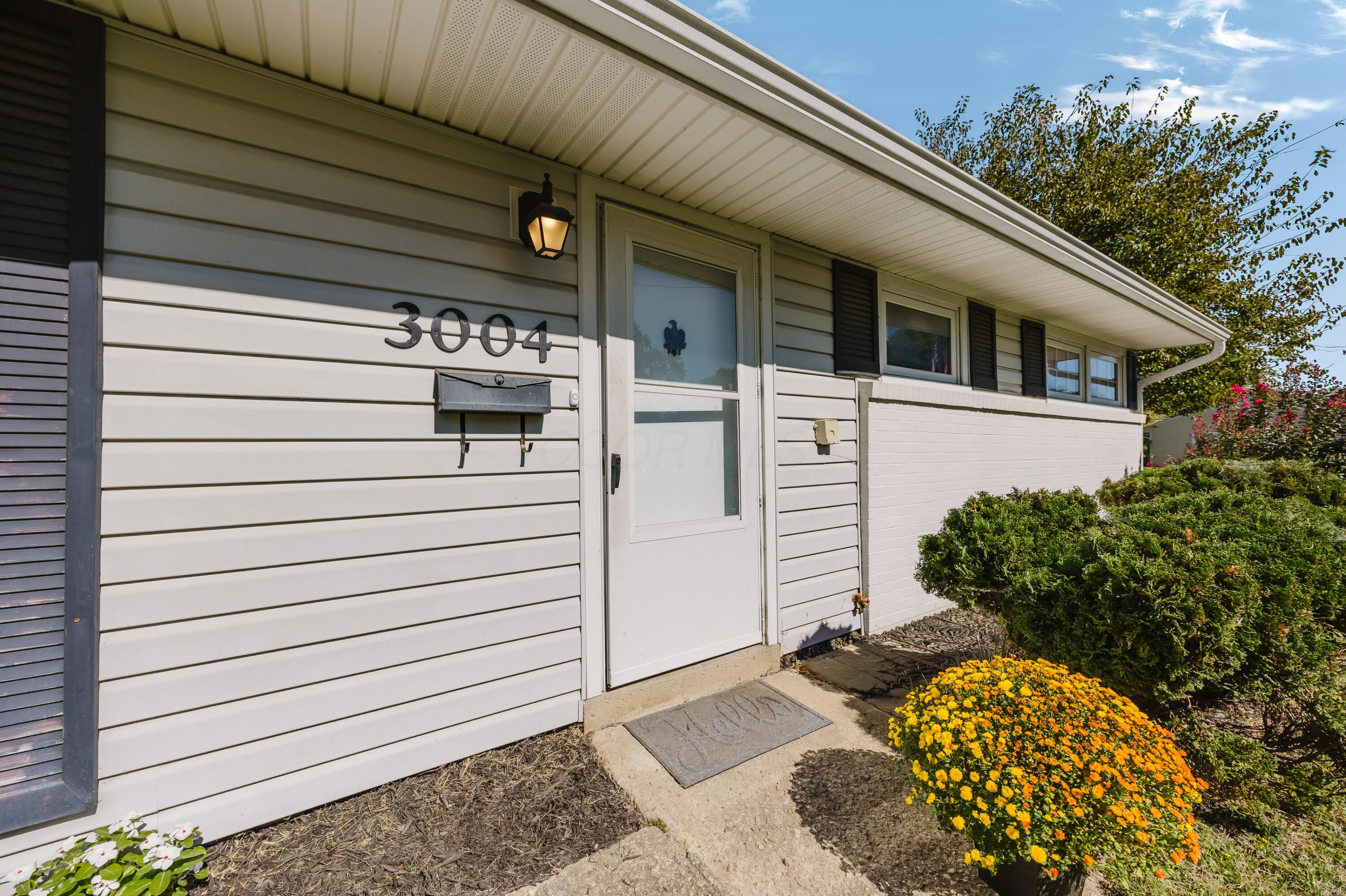Home entrance with 3004 above the mailbox, a white storm door, and bright yellow-orange flowers.
