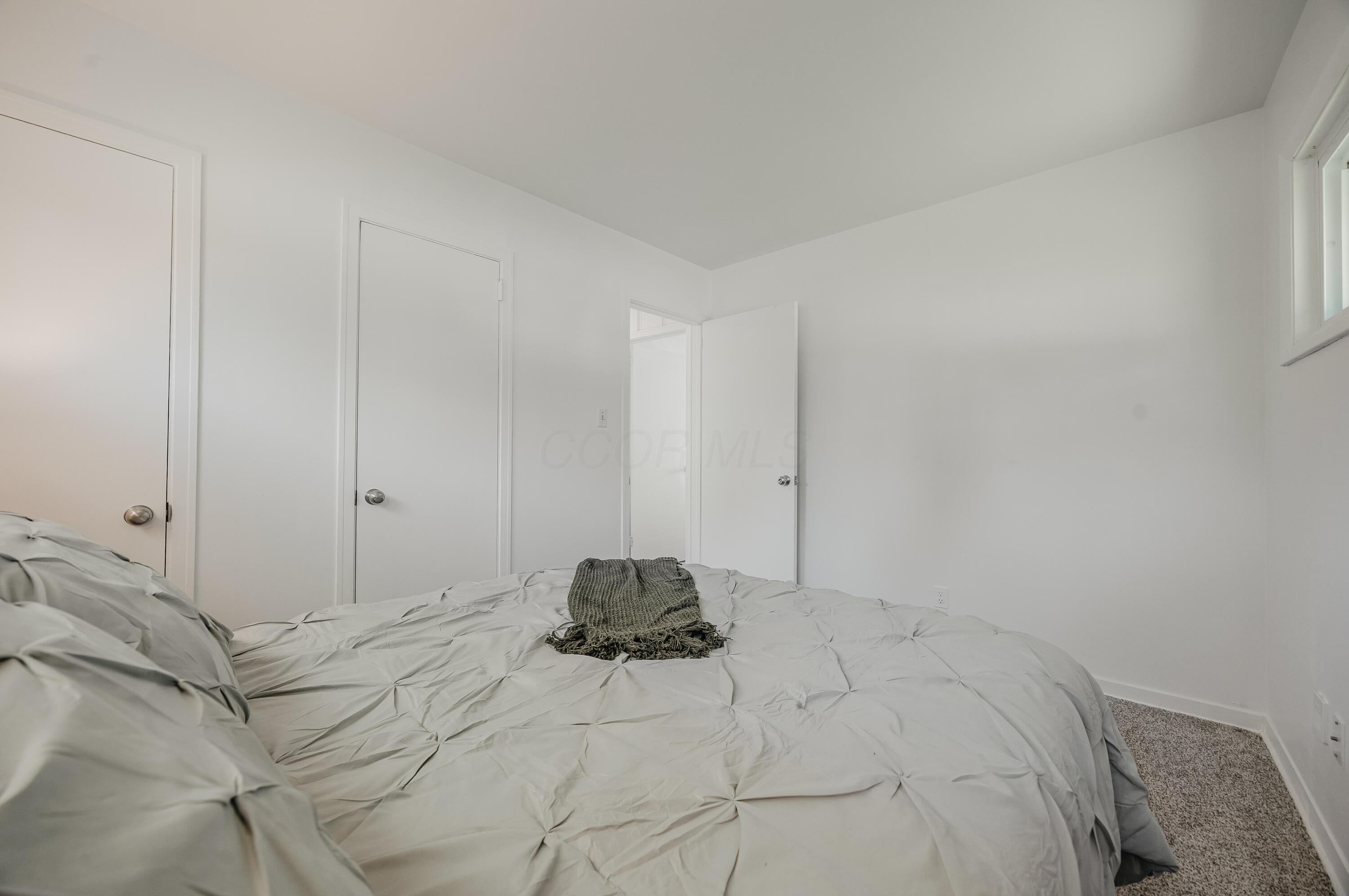 Bright white bedroom with a large pale bed, dark throw, two doors, and a high window.