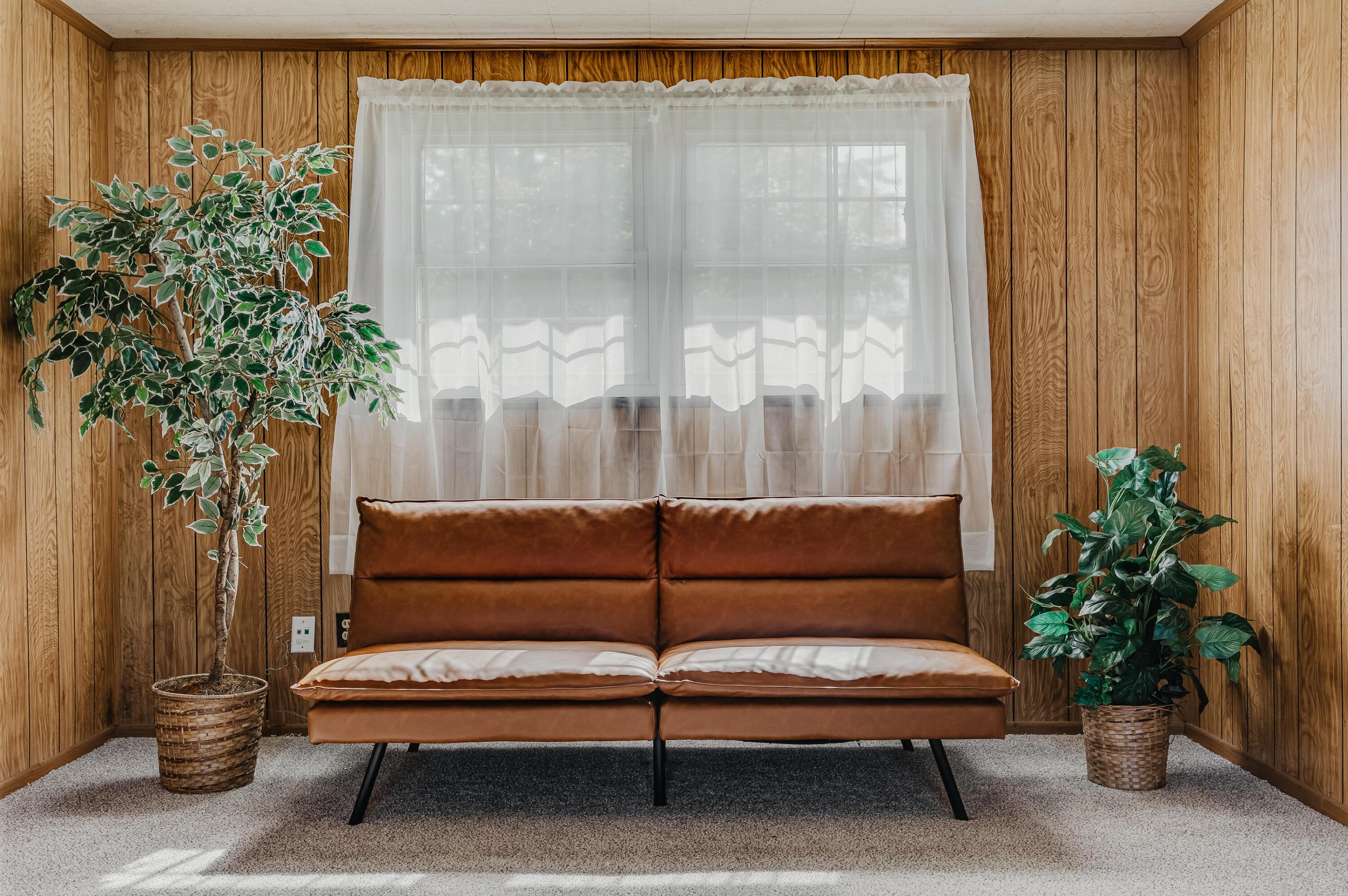Brown sofa beneath a curtained window in a wood-paneled room, flanked by potted plants.
