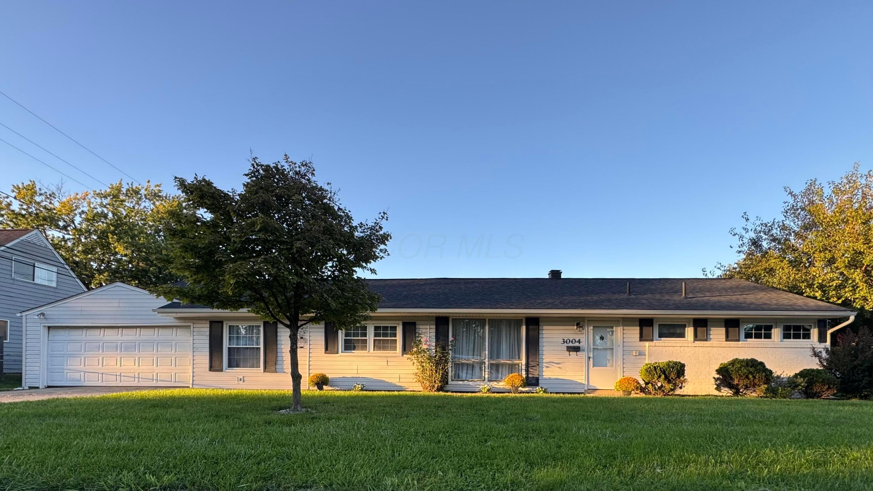 Single-story ranch home with an attached garage, brick facade, and mature trees across the front lawn.