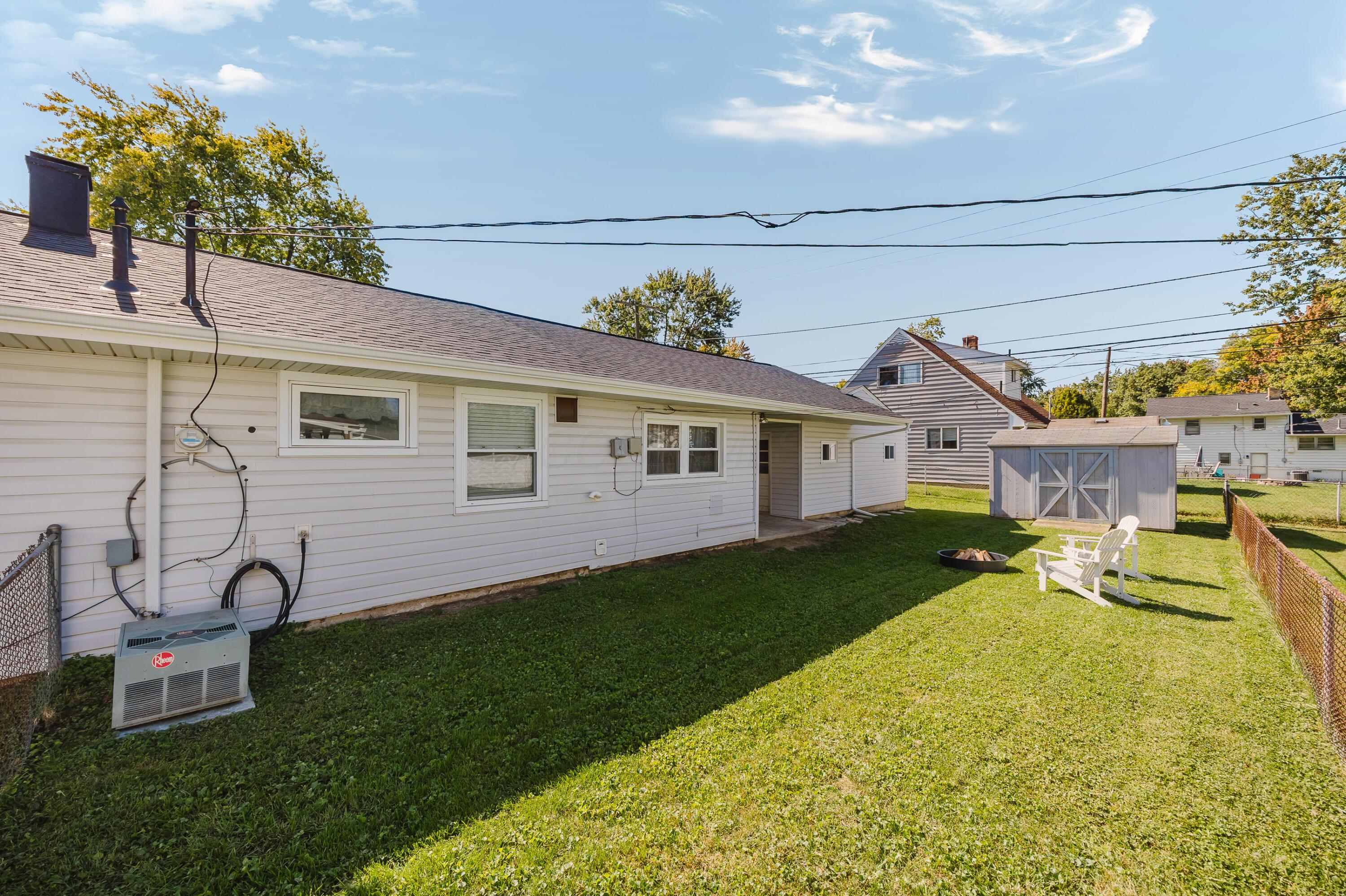 Rear exterior of a single-story home with a fenced lawn, storage shed, and white patio chairs.