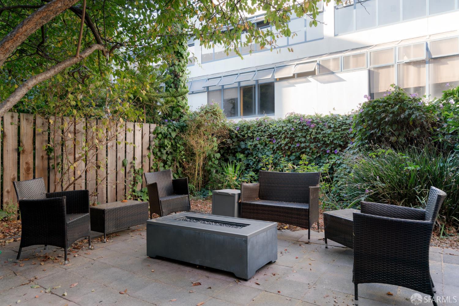 Garden patio with woven seating around a rectangular fire table, bordered by lush planting and a wooden fence.