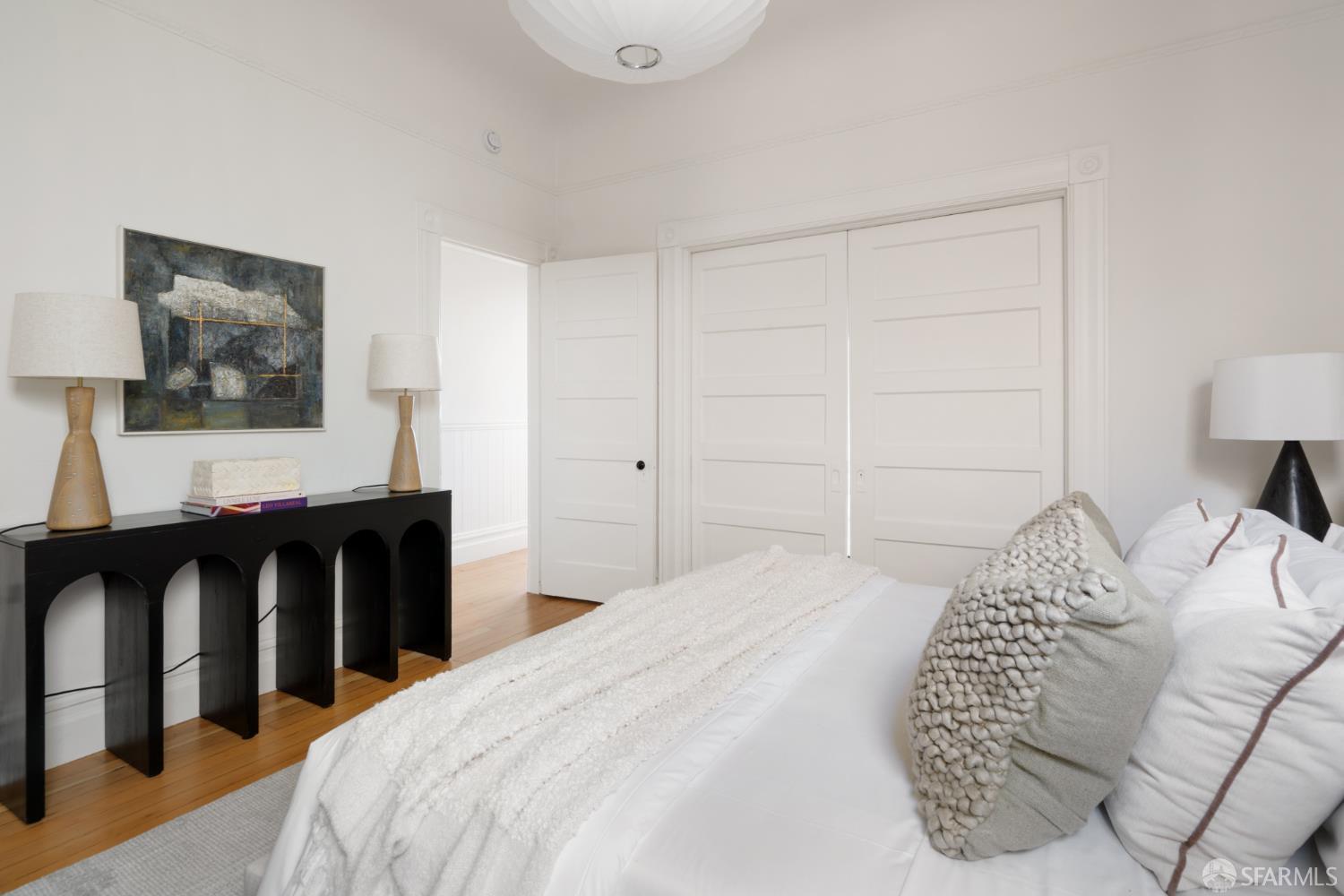 Staged bedroom with a white bed, paneled closet doors, hardwood floors, and a black console table.