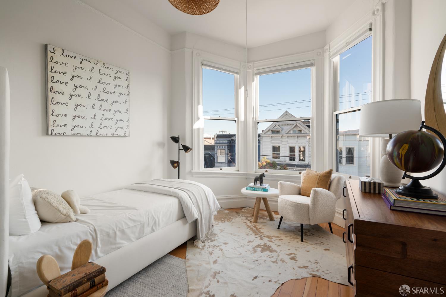 Bright staged bedroom with a white bed, sitting area, and tall bay windows overlooking neighboring houses.
