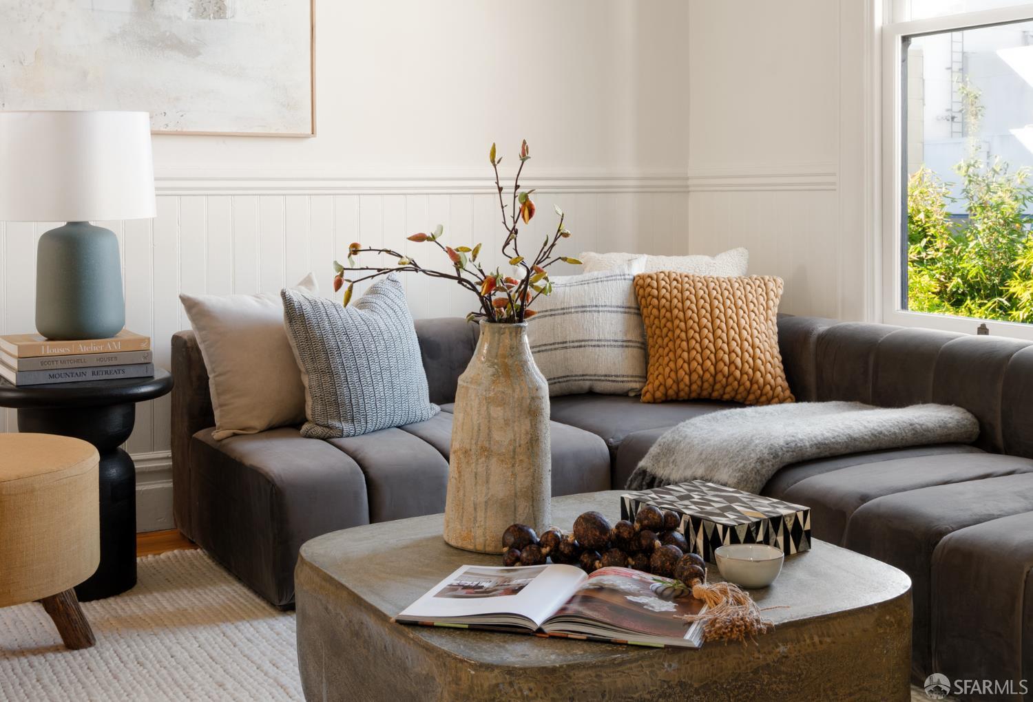 Living room with a gray sectional, textured pillows, and a stone coffee table beside a bright window.