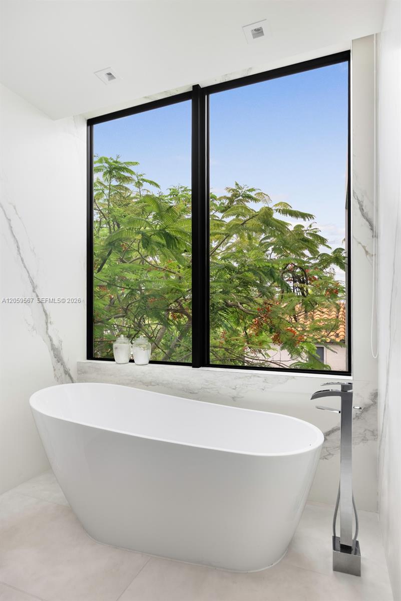 Modern bathroom with a freestanding tub beneath a large window overlooking leafy trees.