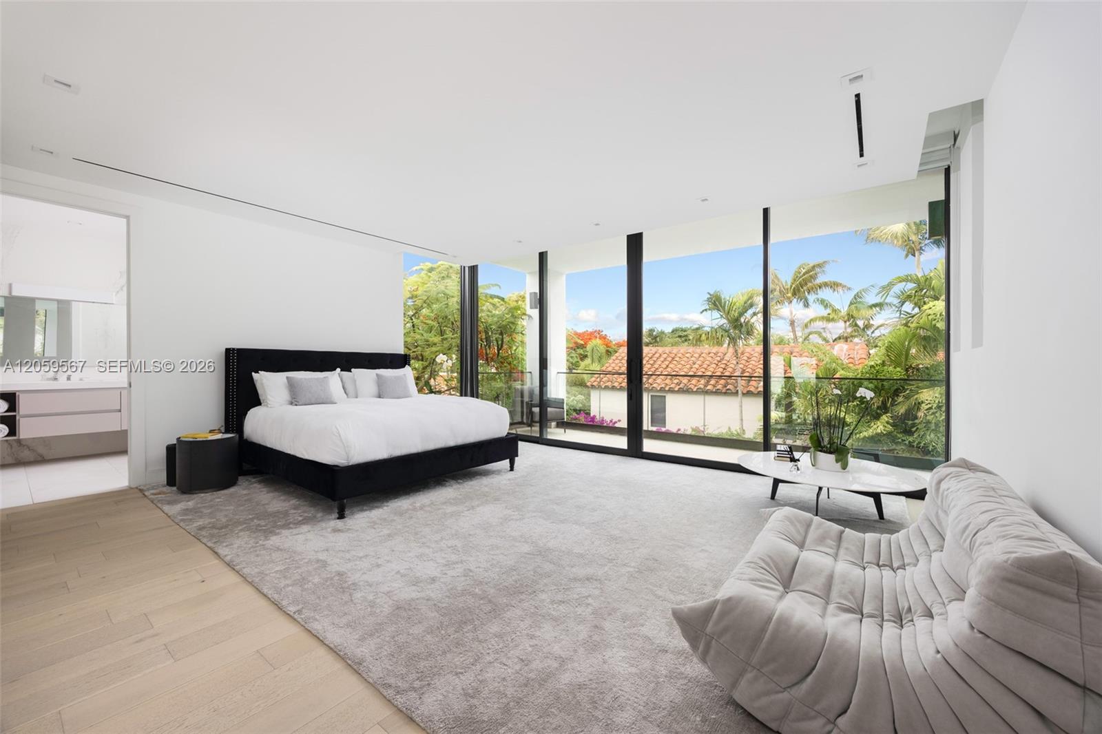 Bright bedroom with floor-to-ceiling windows overlooking palm trees and a tiled roof.