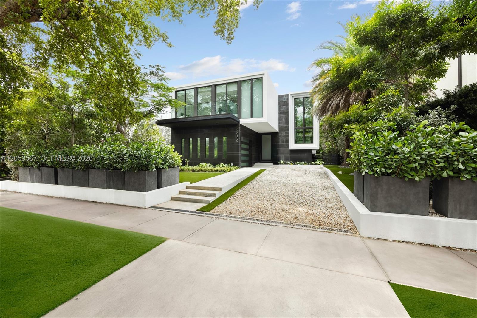 Modern two-story home with large windows, a gravel driveway, and tropical landscaping.