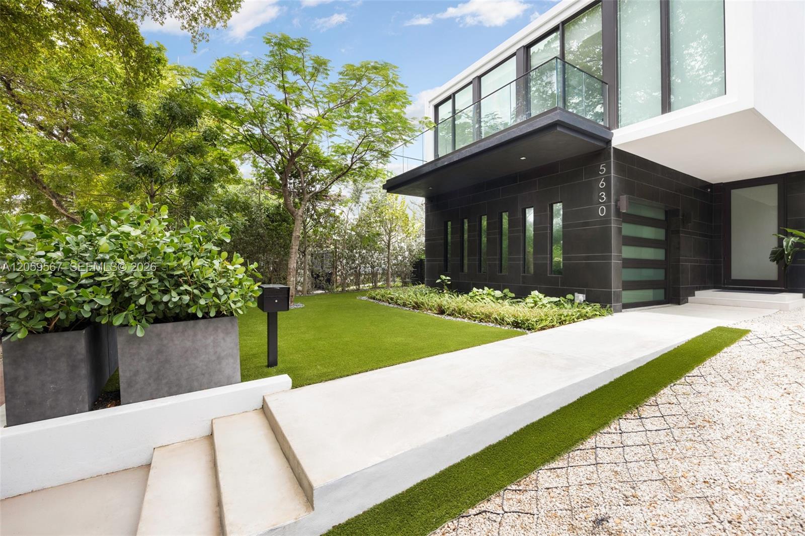 Modern home entrance with black tile facade, glass balcony, landscaped lawn, and white walkway.