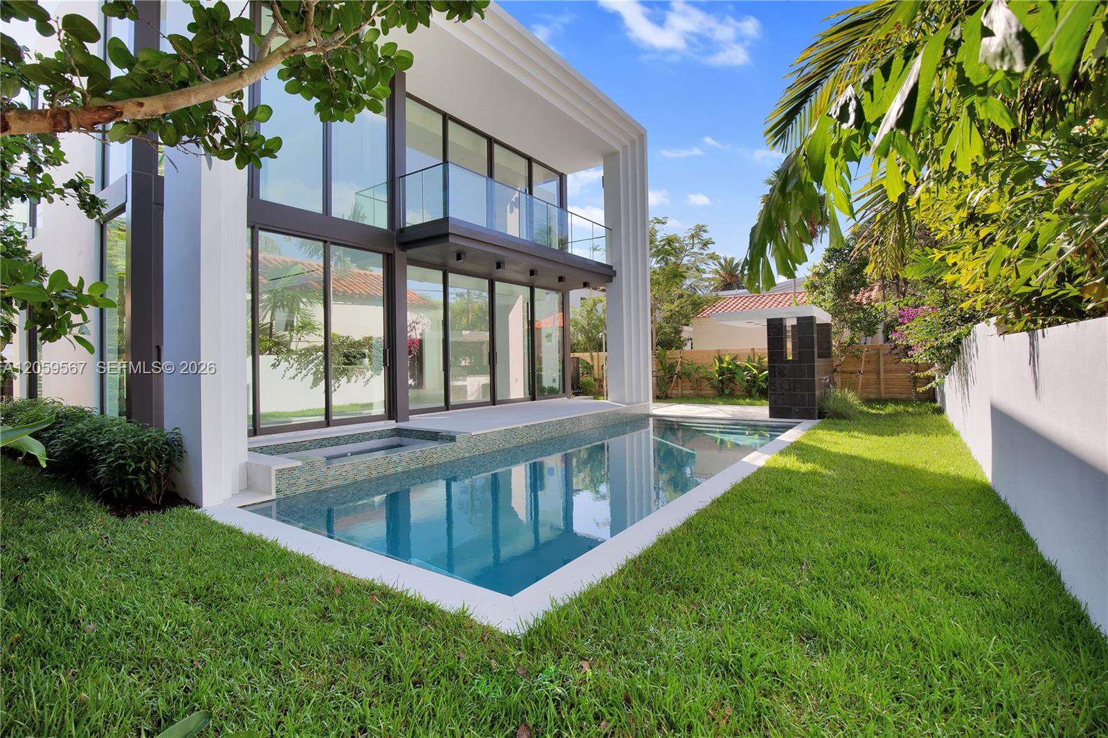 Modern two-story home with a glass balcony overlooking a narrow backyard pool and tropical landscaping.