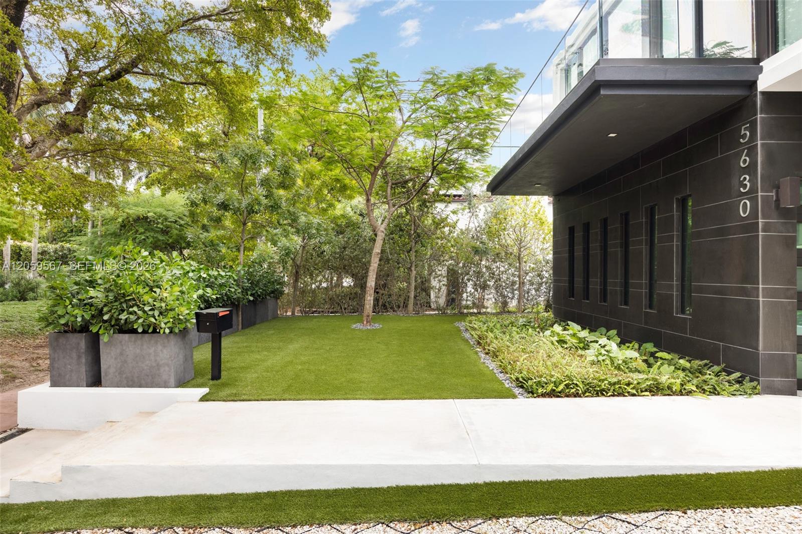 Modern home entrance with dark tile, a manicured lawn, raised planters, and mature trees.