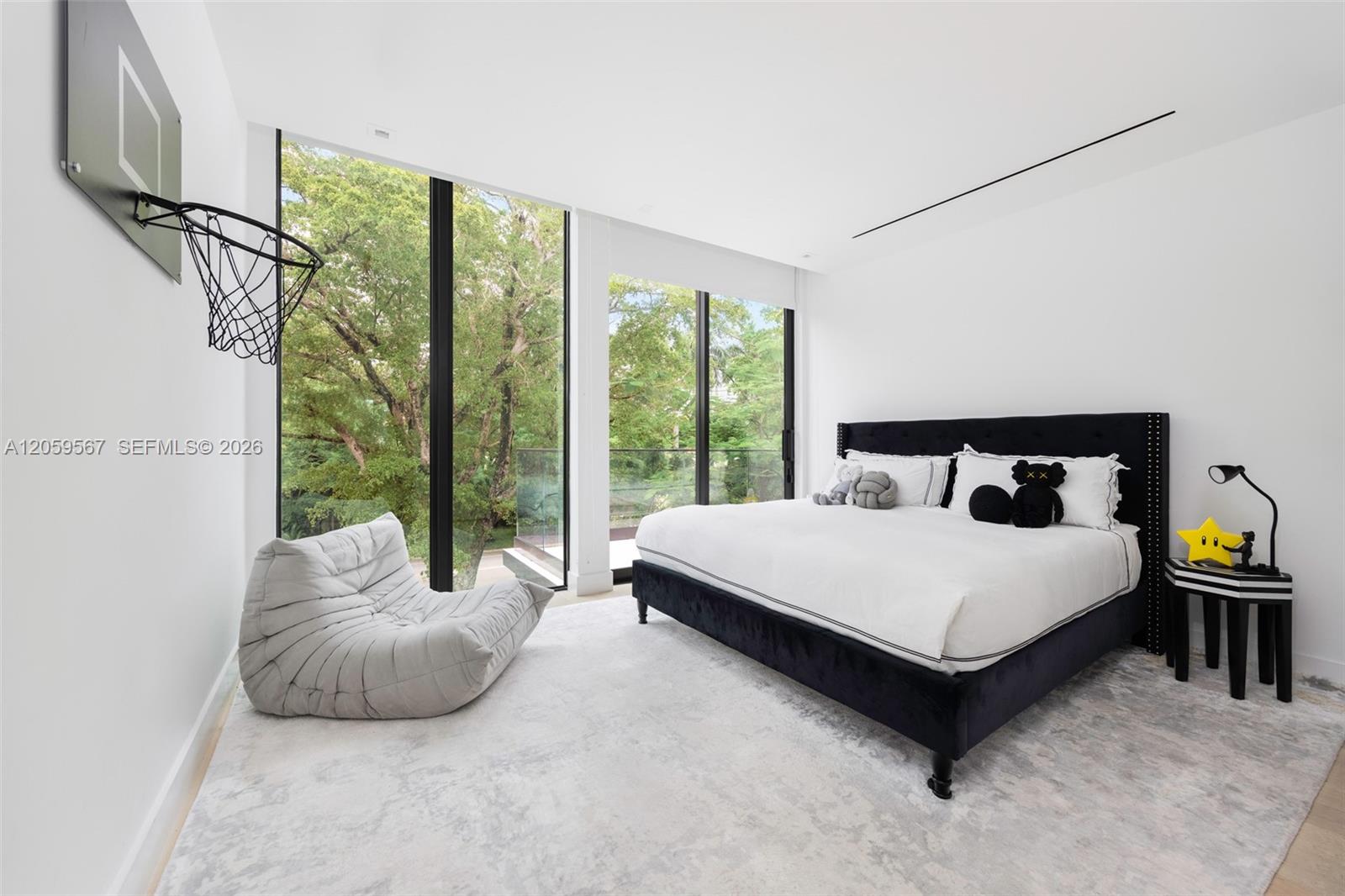 Bedroom with a black bed, wall-mounted basketball hoop, and tall windows overlooking leafy trees.