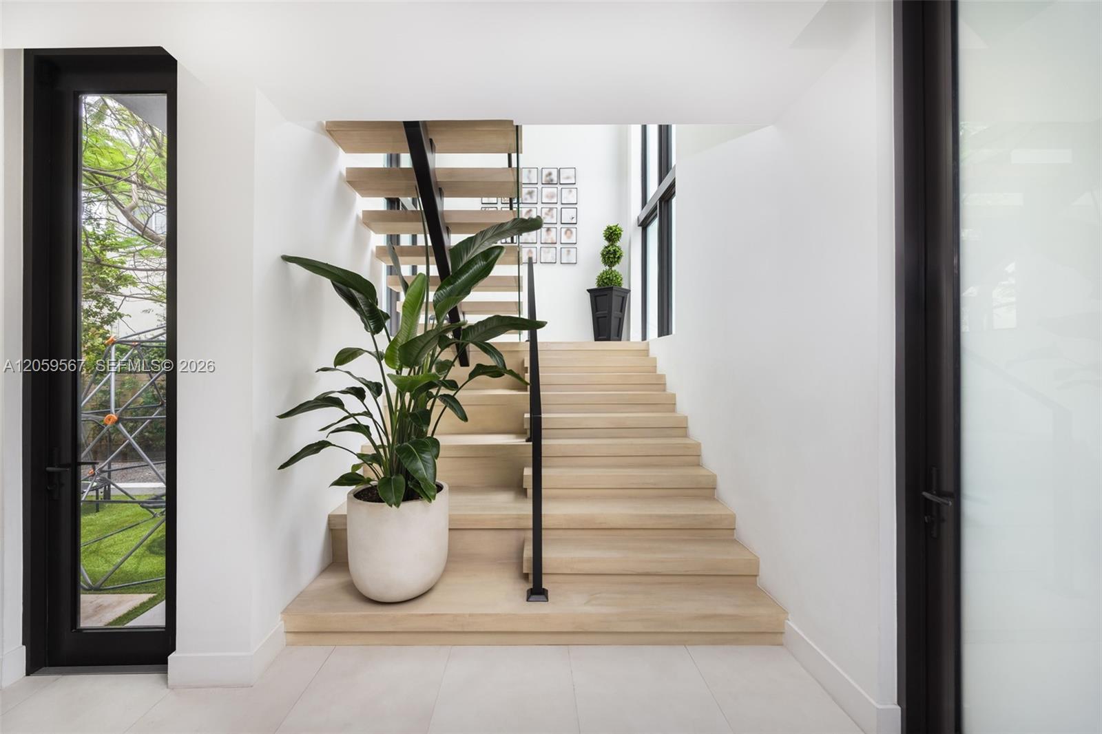 Light wood staircase with a black railing, tall windows, and a potted tropical plant.