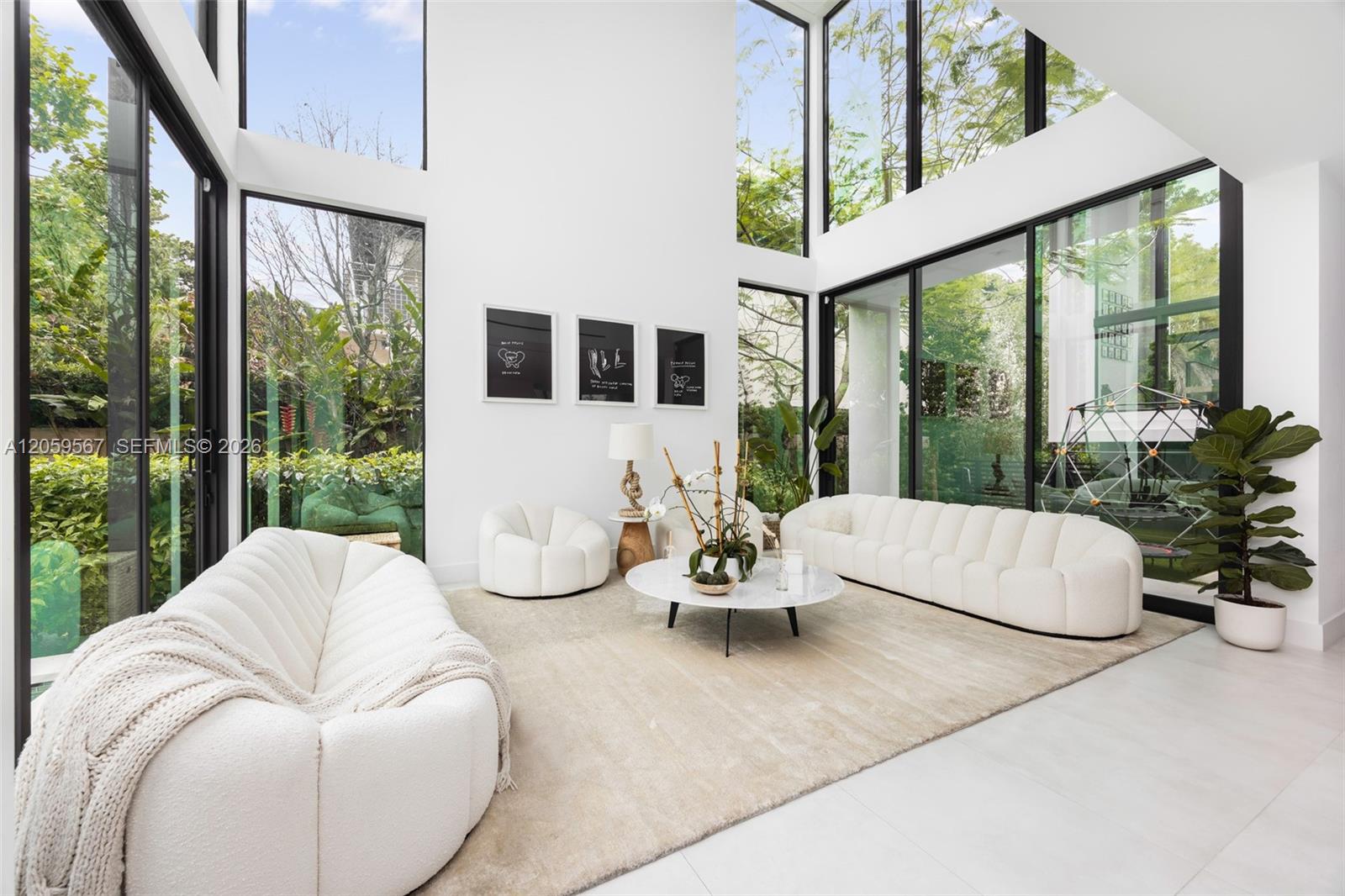 Double-height living room with white sofas and expansive black-framed windows overlooking greenery.