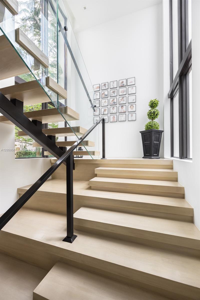 Interior staircase with broad pale wood steps, glass railing, tall windows, and a potted topiary on the landing.
