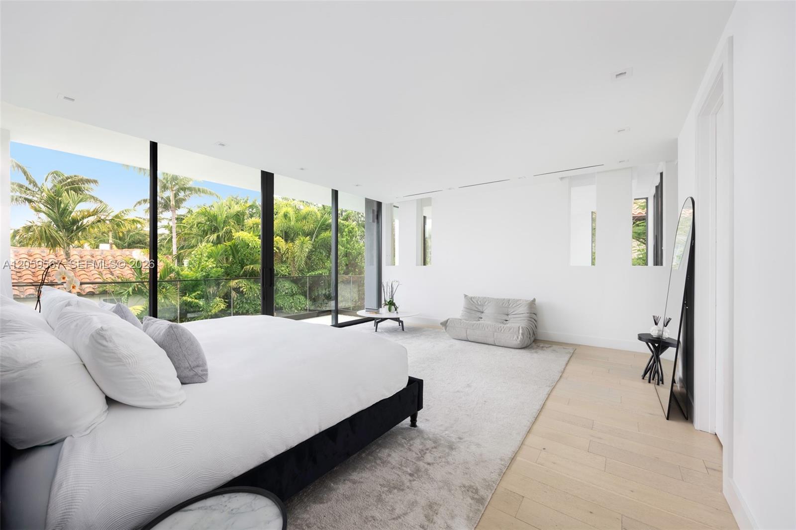 Modern bedroom with a large white bed and floor-to-ceiling windows overlooking palms.