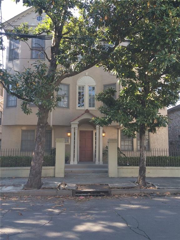 Beige stucco home with a red front door, arched portico, iron fence, and mature trees.
