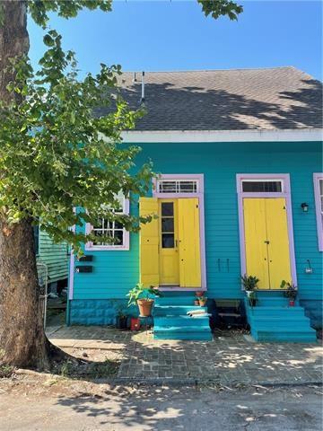 Turquoise house with yellow doors and pink trim, shaded by a large tree.