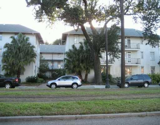 Multi-story residential building with balconies, mature trees, and cars parked along the curb.