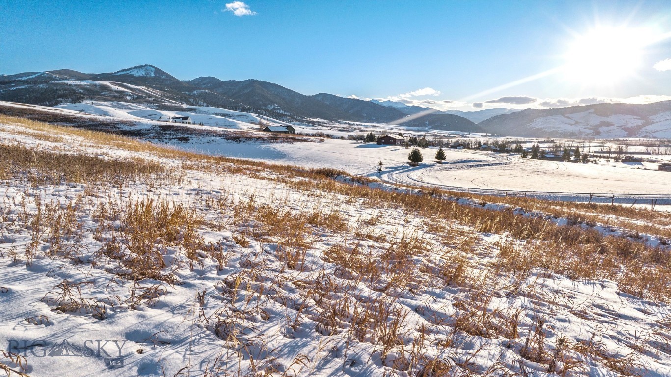 Snow-covered vacant lot with dry grasses, scattered homes and forested mountains in the distance.