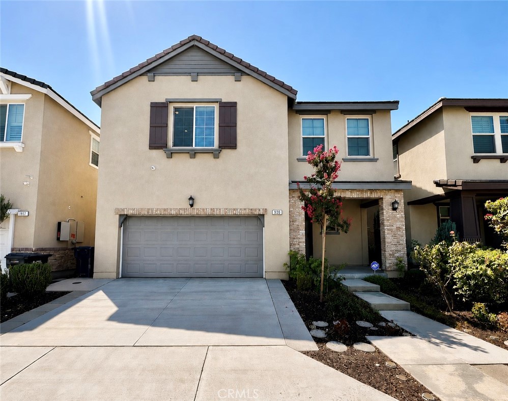 Two-story beige stucco home with a two-car garage, dark shutters, and landscaped entry.
