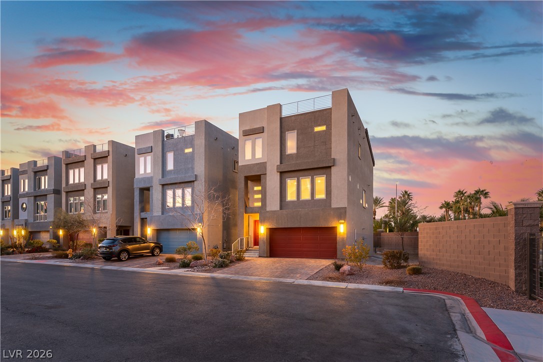 Modern three-story townhome with a dark red garage door beneath a pink sunset sky.