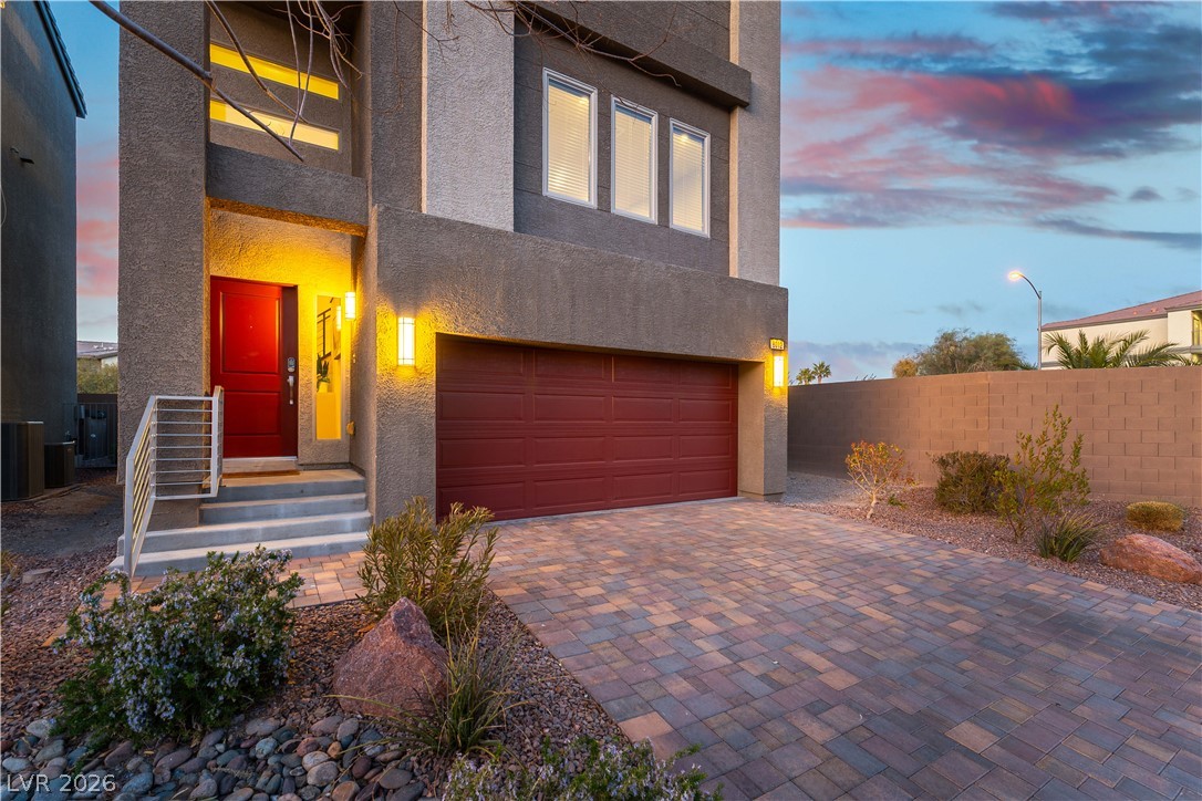 Two-story gray stucco home with a red entry door, double garage, and paver driveway at dusk.