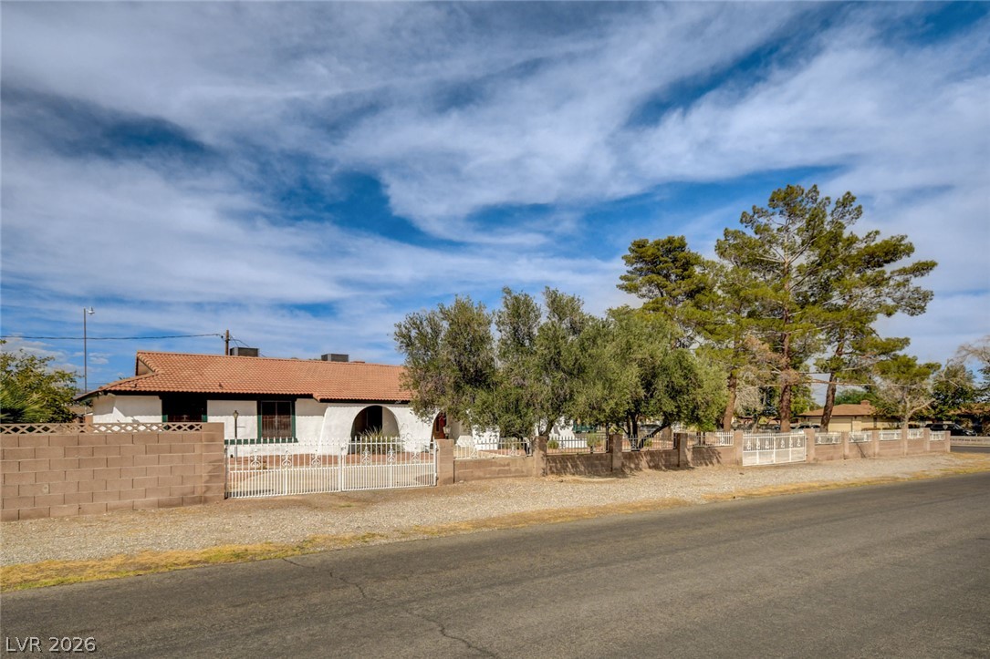 Single-story stucco home with a tiled roof, arched entry, white gate, and mature trees along the street.