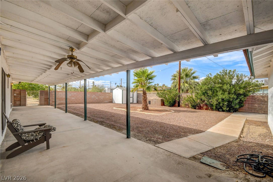 Covered backyard patio with a ceiling fan overlooking a gravel yard, palm trees, and a block wall.