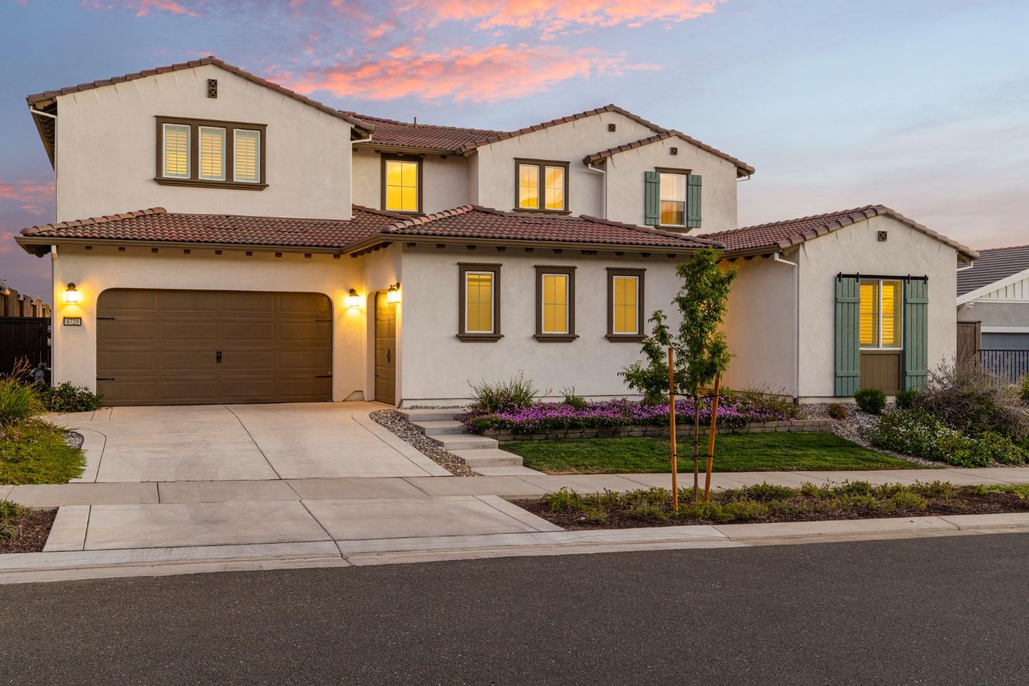 Two-story stucco home with a red tile roof, green shutters, and a landscaped front yard at dusk.