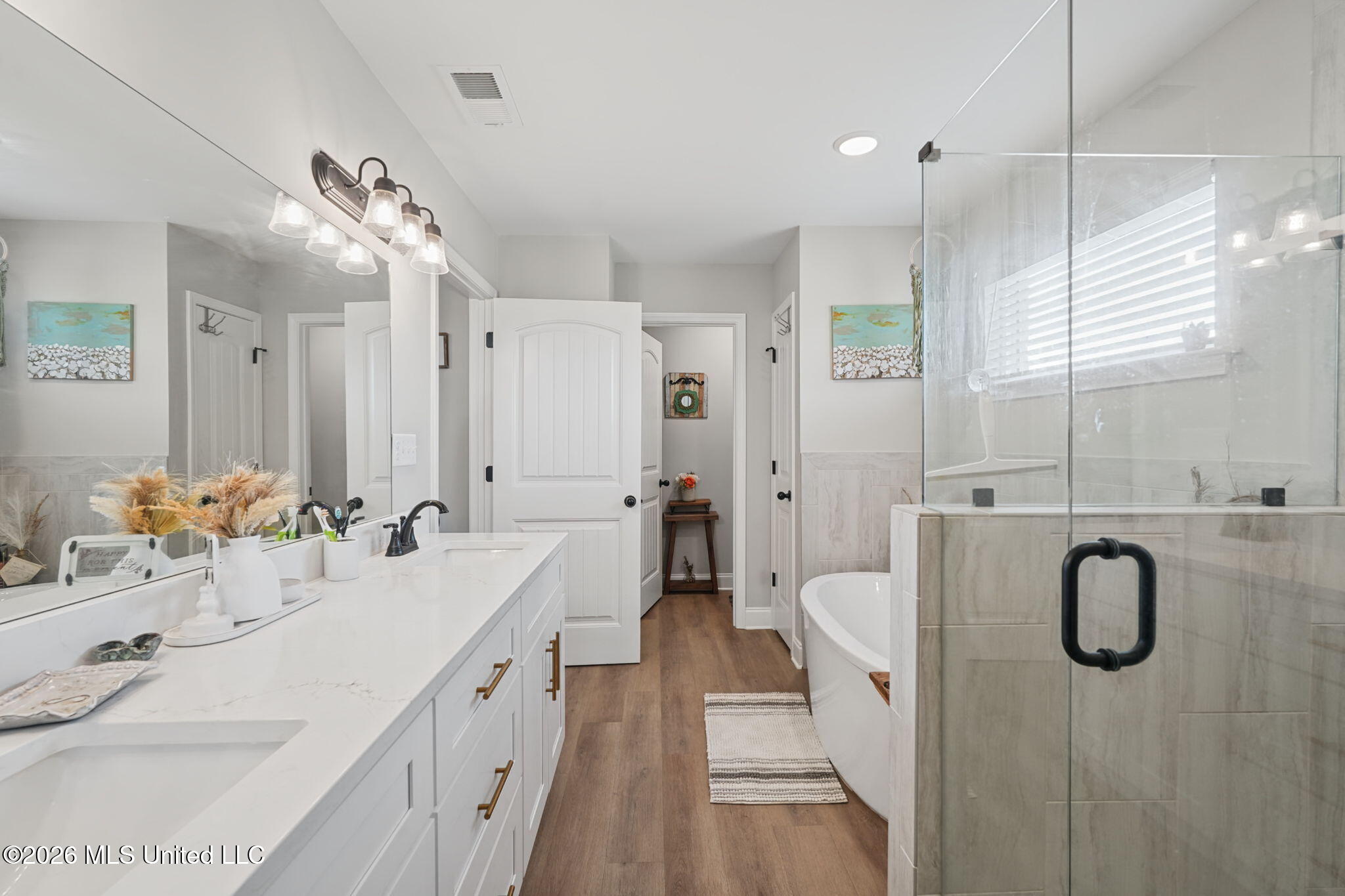 Bright bathroom with a long white vanity, glass-enclosed shower, freestanding tub, and wood-look flooring.