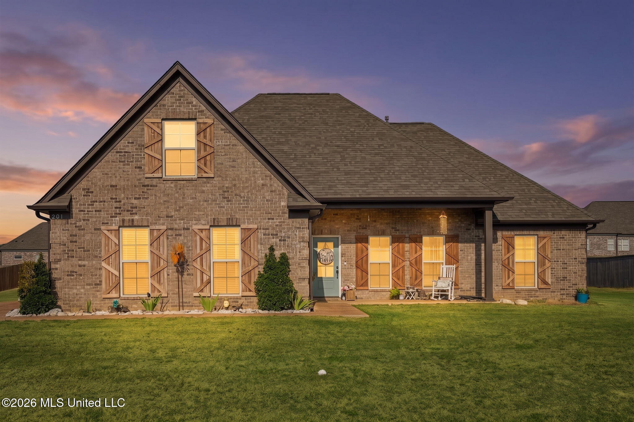 Brick house with wood shutters and warmly lit windows at dusk, viewed across a broad lawn.