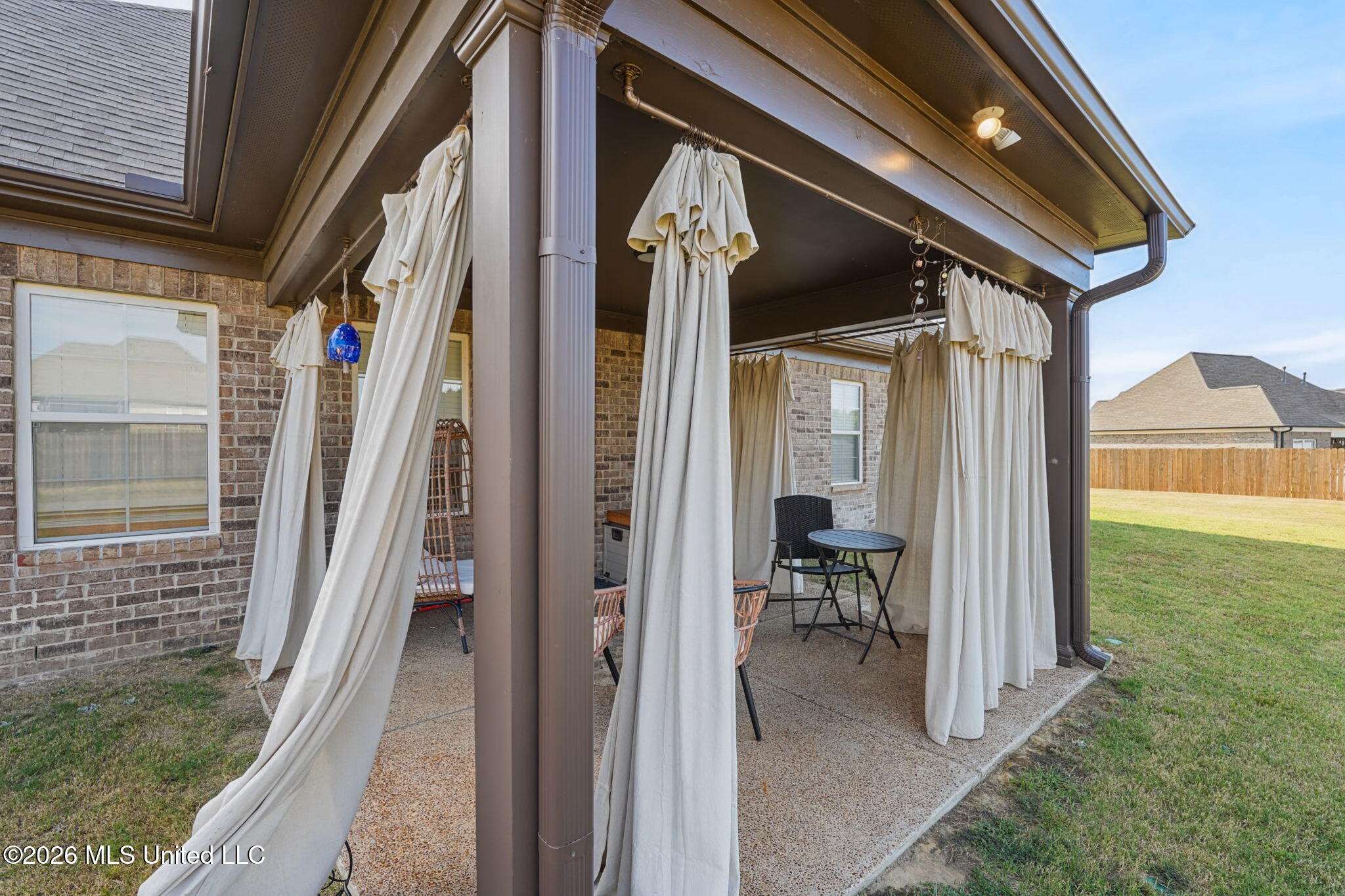 Covered brick patio with white curtains, outdoor seating, and a fenced backyard.