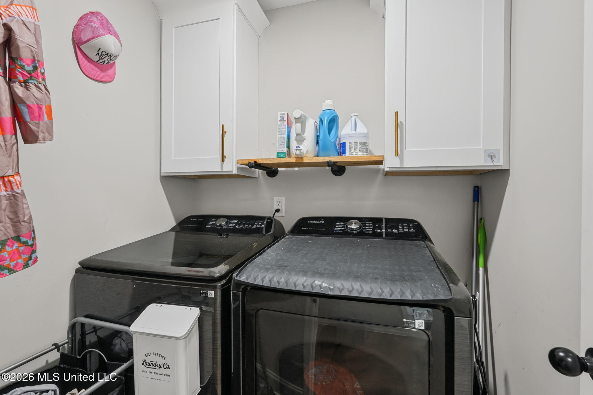Laundry room with dark washer and dryer beneath white cabinets and a shelf of detergent.