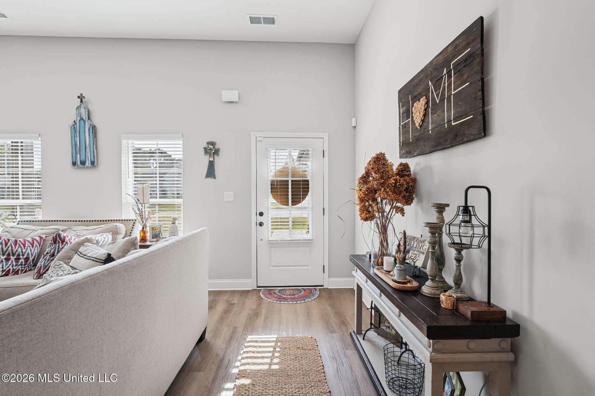 Living room and entryway with a white sofa, bright front door, and dark console table beneath a HOME sign.