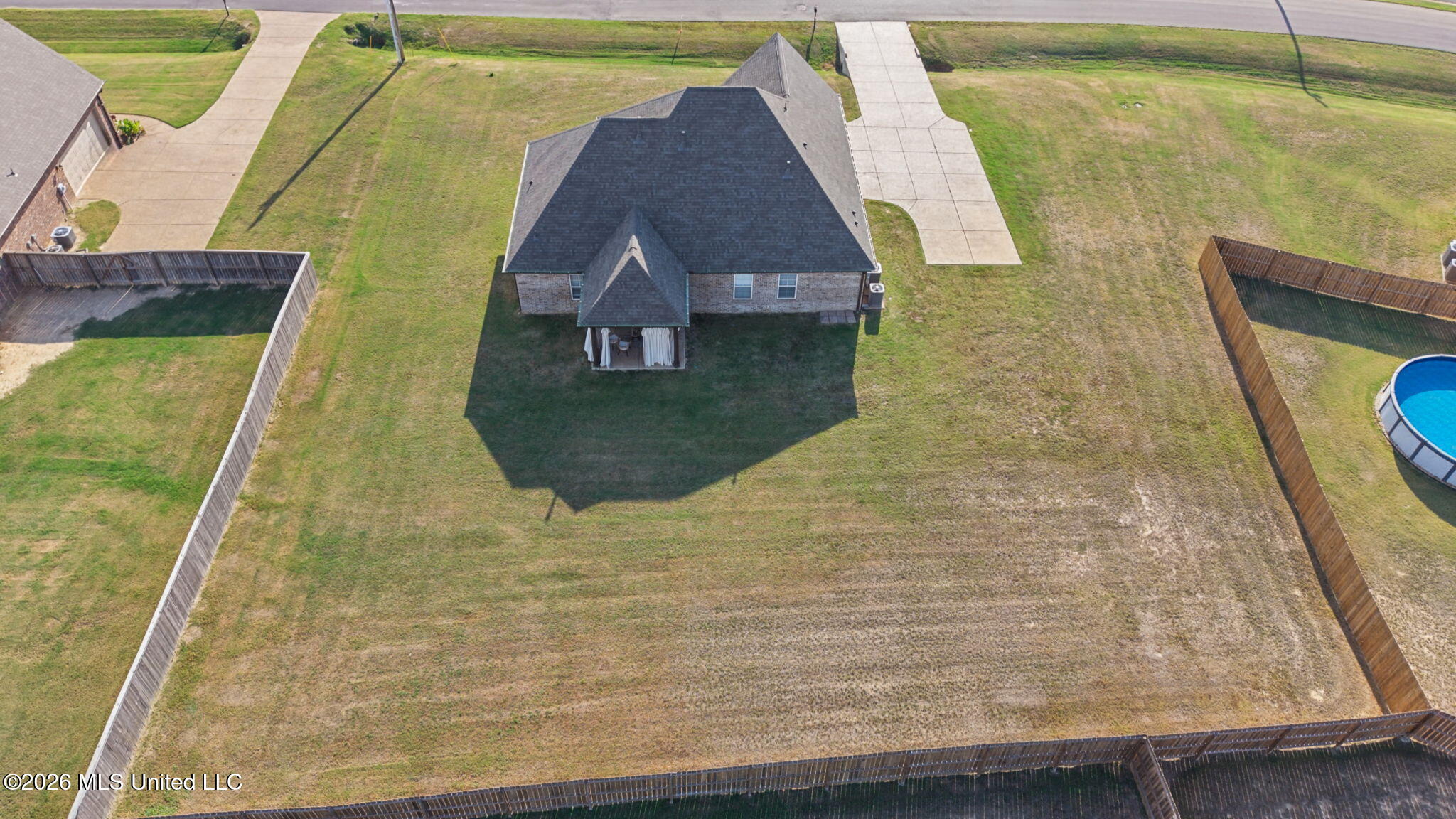 Aerial view of a brick house and spacious fenced backyard, with a covered patio and an above-ground pool partly visible nearby.