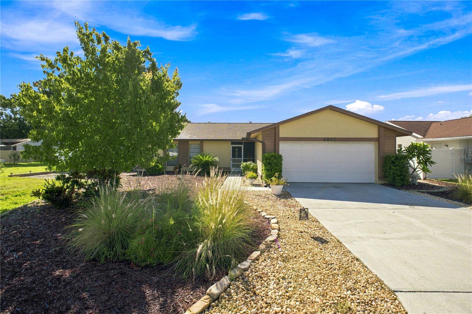 Single-story home with an attached two-car garage, landscaped stone beds, and a large shade tree.
