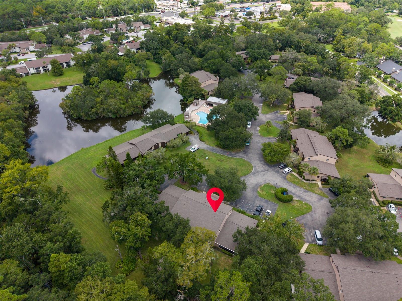 Aerial view of a wooded residential community with a pond, pool, and red map pin marking a building.