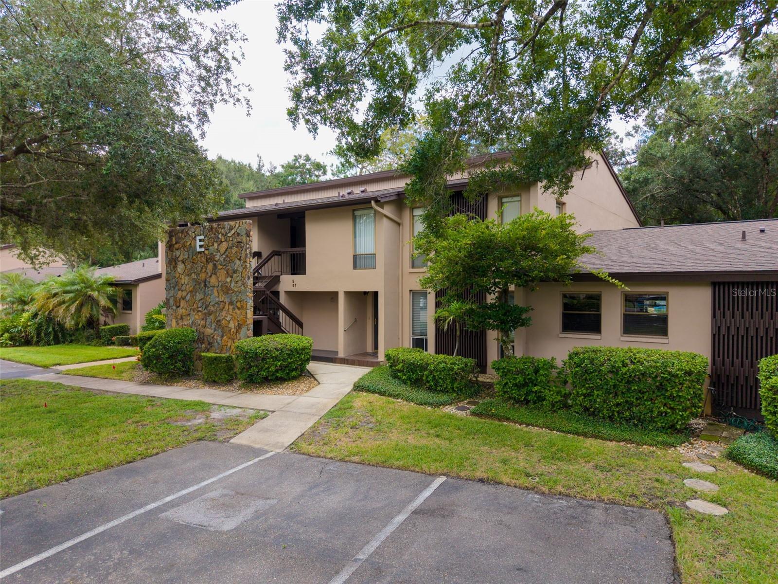 Two-story beige condominium framed by mature trees, with a stone entrance marker labeled E and parking in front.