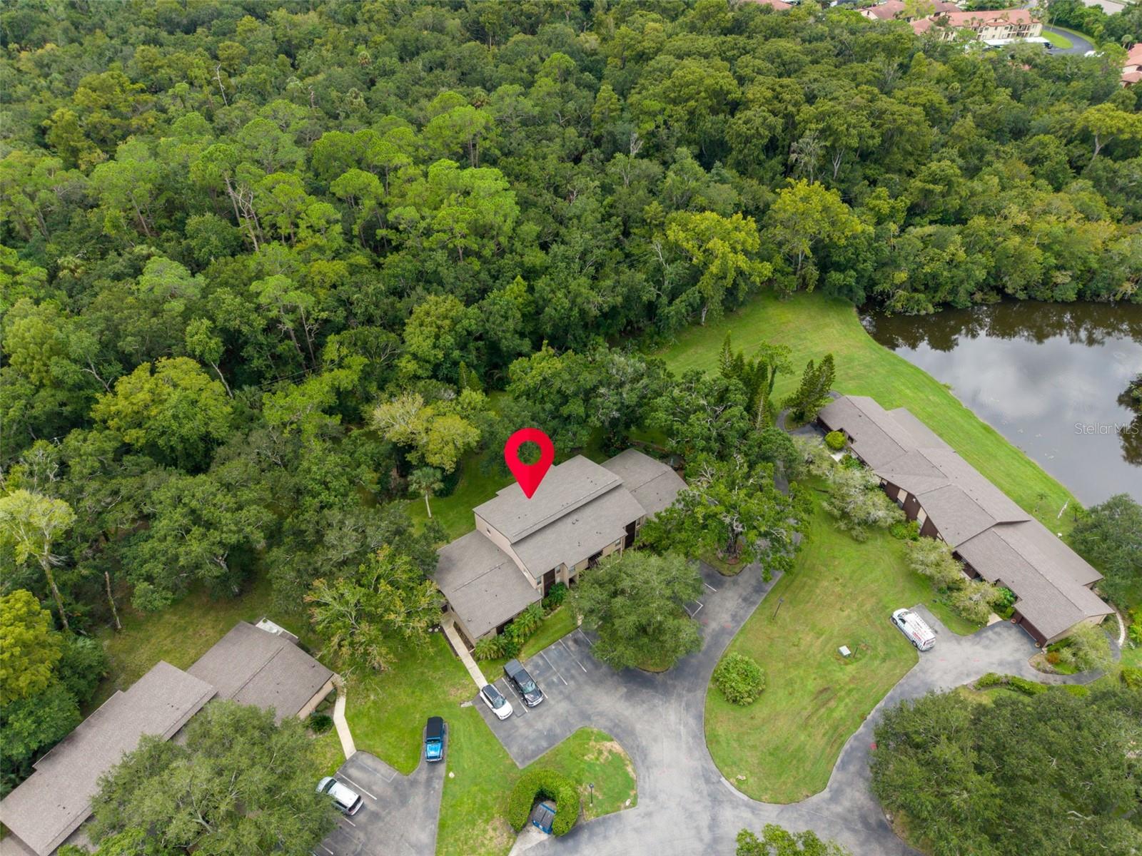 Aerial view of a condo building marked with a red pin beside wooded grounds and a pond.