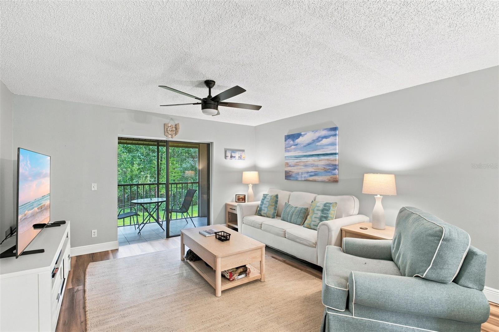 Living room with light seating and a sliding glass door opening to a greenery-framed balcony.