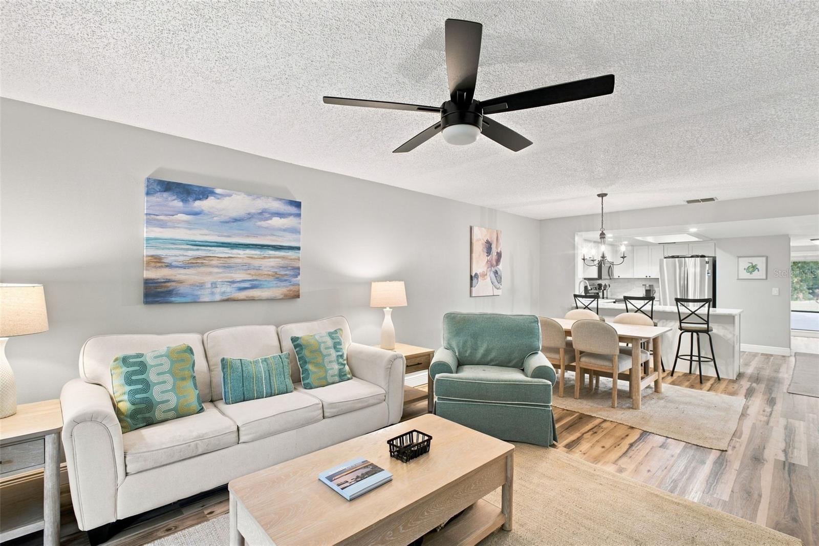 Furnished living room opens to a dining area and kitchen, with a black ceiling fan.